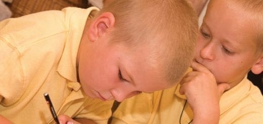 Two young boys in yellow shirts, one writing and the other whispering.