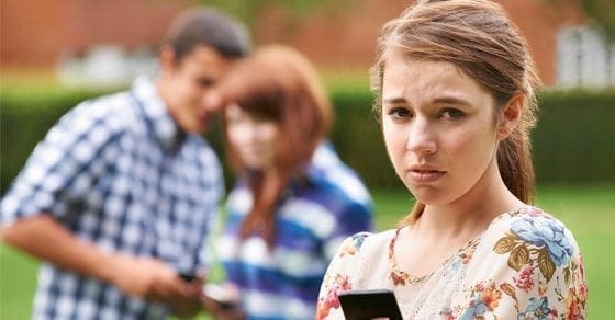 A young girl looks worriedly at her phone while two teenagers whisper behind her.
