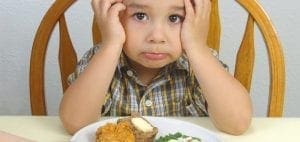 Child with hands on head looking at a plate of food.