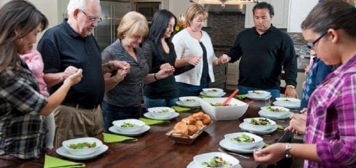 Family with hands clasped in prayer around a dining table.