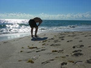 Person bending over on a sandy beach with the ocean in the background.