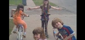 Four children are outside on a sidewalk with bikes and scooters.