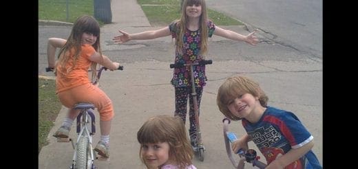 Four children are outside on a sidewalk with bikes and scooters.