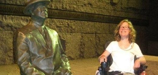 A woman in a wheelchair smiles next to a statue of a man in a hat.