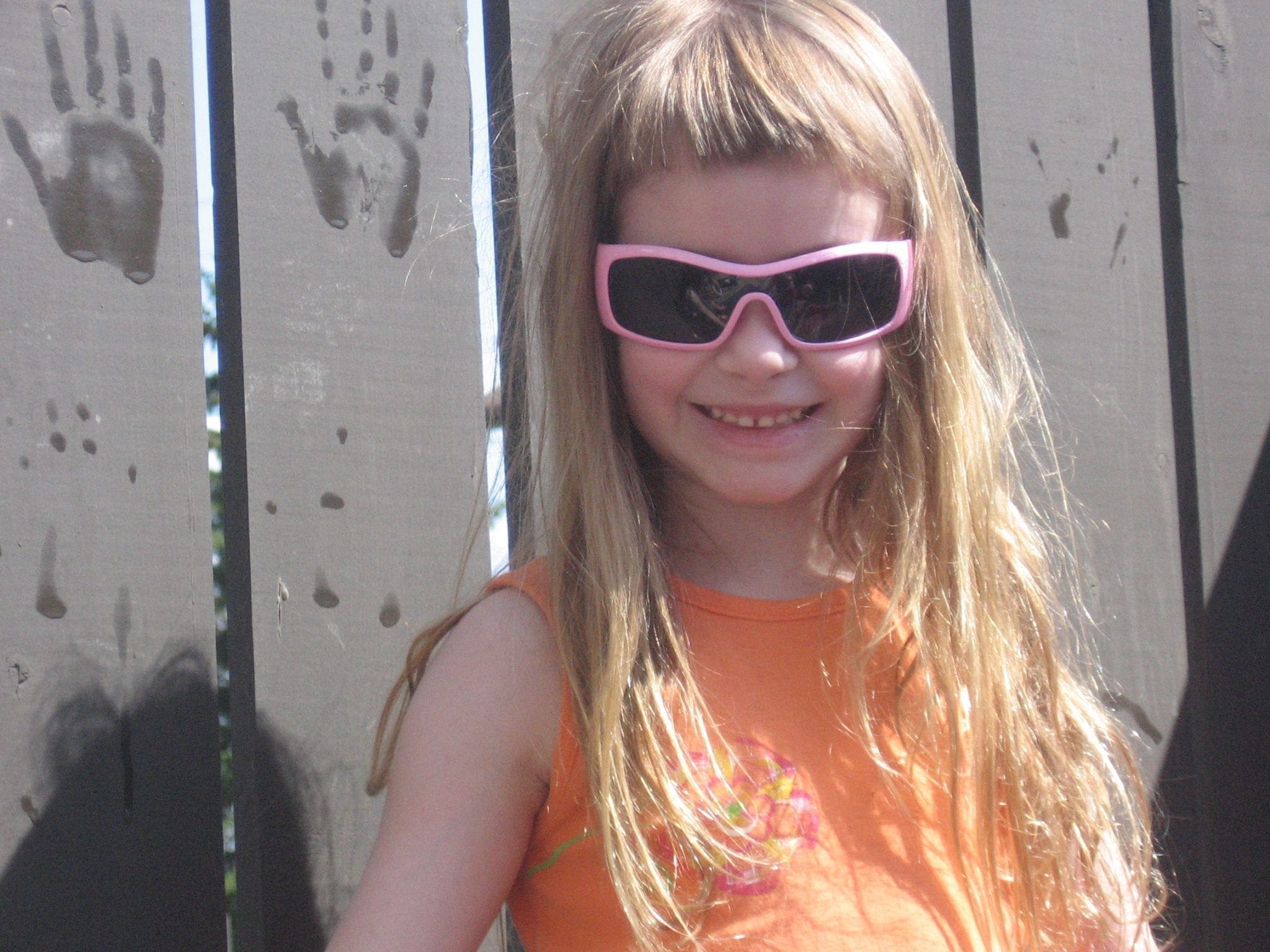 A young girl with long blond hair and sunglasses smiles in front of a wooden fence.