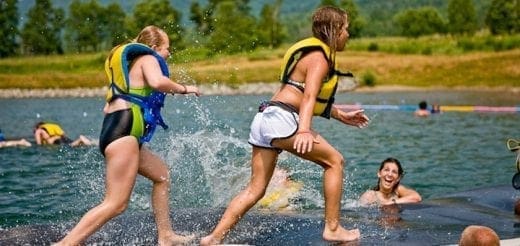 Two young women in life jackets run towards the water, splashing.