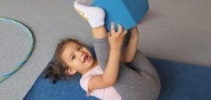 A young child lies on a blue mat and holds a blue block above their legs.