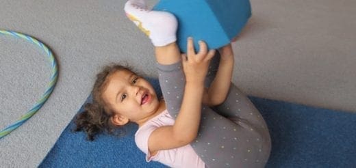 A young child lies on a blue mat and holds a blue block above their legs.