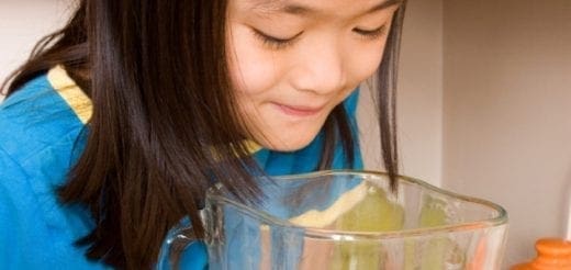 A girl in a blue shirt leans over a blender full of green smoothie.