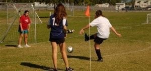 Three girls play soccer on a grassy field near a goal.