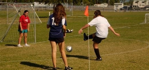 Three girls play soccer on a grassy field near a goal.