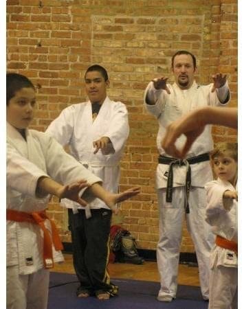 Two men and two children in martial arts uniforms practice stances in front of a brick wall.