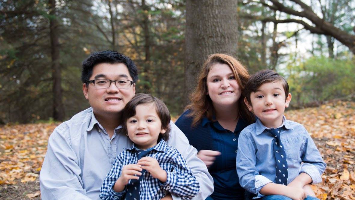 A family of four poses for a photo outdoors with autumn leaves on the ground.