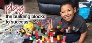 Boy smiling and giving a peace sign near building blocks.