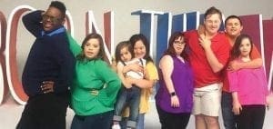 A group of eight people stand in front of a large sign that says "BORN THIS WAY.