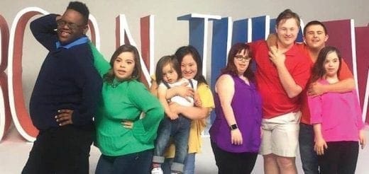 A group of eight people stand in front of a large sign that says "BORN THIS WAY.
