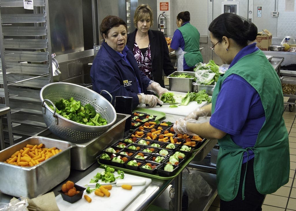 Three people in a kitchen preparing food, including chopped vegetables and a tray of food portions.