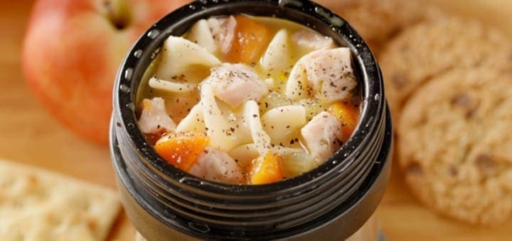 Chicken noodle soup in a black container with an apple and crackers.