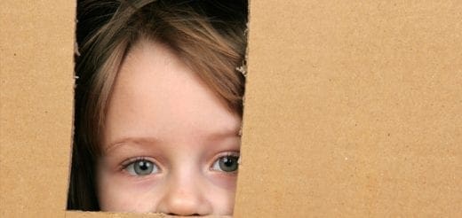 Close-up of a young child's face peering through a gap in a cardboard box.