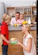 A mother hands a lunchbox to her son, with his father and sister smiling in the background.