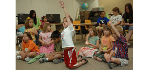 Children sit on the floor in a classroom, with one boy kneeling and raising his hand.