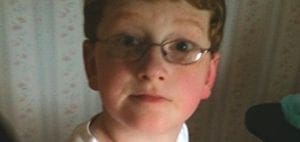 A young boy with red hair and glasses looks directly at the camera.