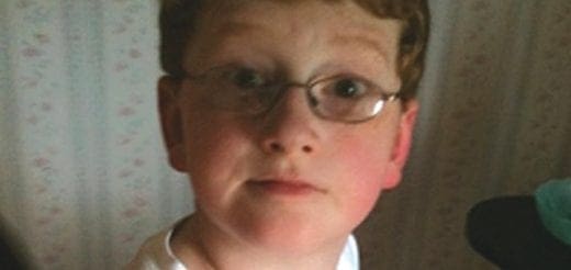 A young boy with red hair and glasses looks directly at the camera.