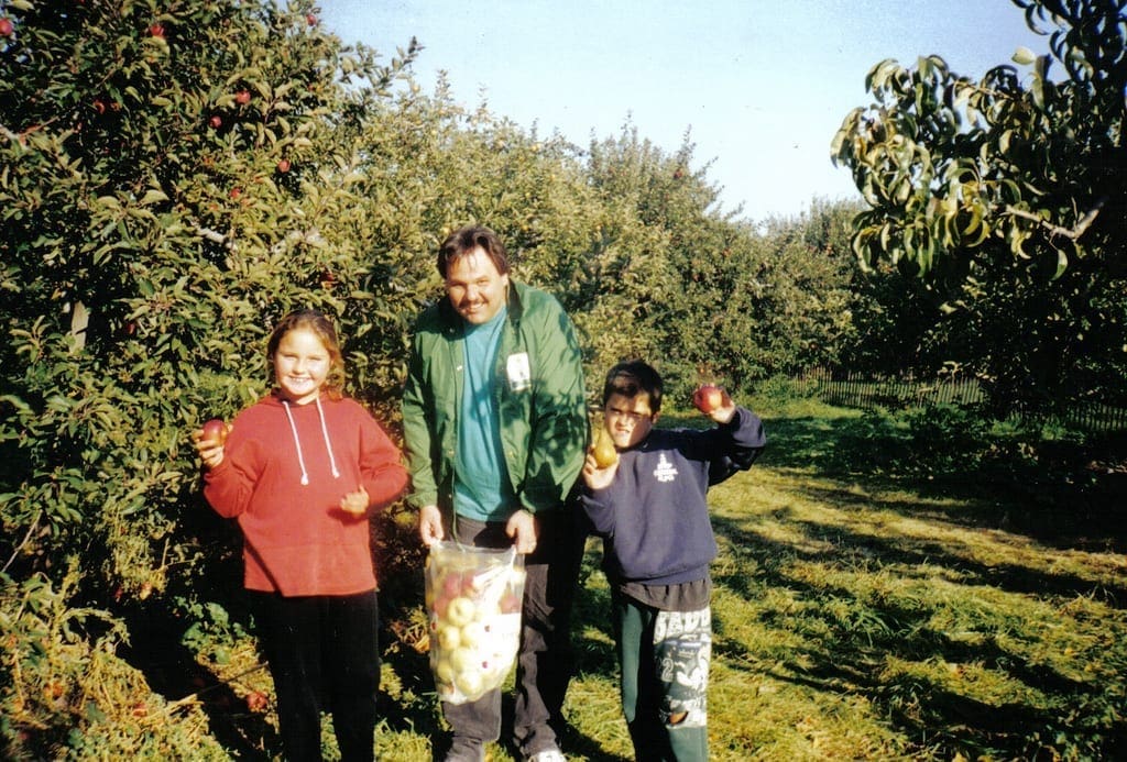 Man holding a bag of apples with two children in an orchard.