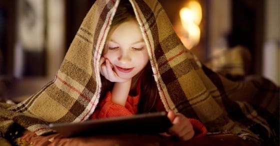 A young girl under a blanket looks at a tablet.