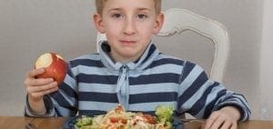 Young boy in a striped shirt holding an apple while sitting at a table with a plate of pasta.