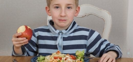 Young boy in a striped shirt holding an apple while sitting at a table with a plate of pasta.