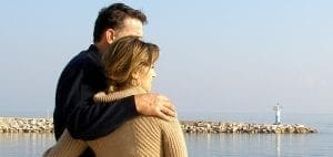 Couple embracing by the sea with a lighthouse in the distance.
