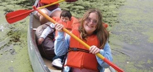 A young woman with long brown hair and a life jacket smiles while holding a canoe paddle.