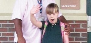 Young girl with Down syndrome waves, standing with an adult.