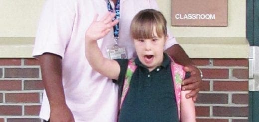 Young girl with Down syndrome waves, standing with an adult.
