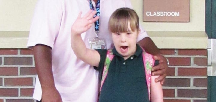 Young girl with Down syndrome waves, standing with an adult.