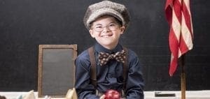 Wade Michael Strite smiles wearing a cap, suspenders, and bow tie in front of a chalkboard.