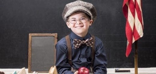 Wade Michael Strite smiles wearing a cap, suspenders, and bow tie in front of a chalkboard.