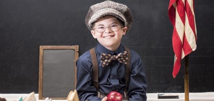Wade Michael Strite smiles wearing a cap, suspenders, and bow tie in front of a chalkboard.