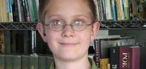 A child with glasses smiles in front of a bookshelf.