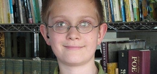 A child with glasses smiles in front of a bookshelf.