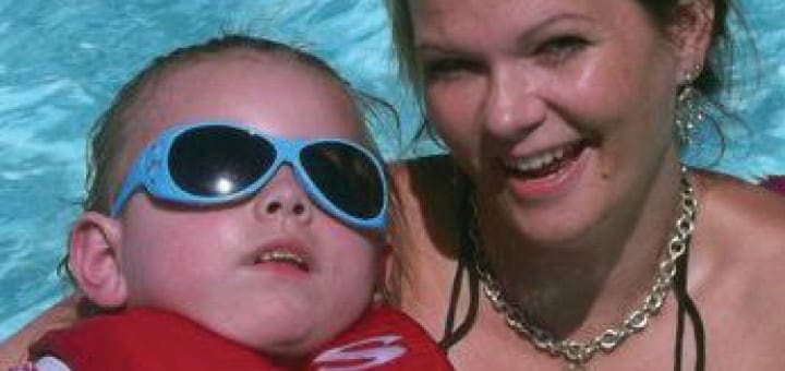 A child wearing sunglasses and a life vest poses with a smiling woman in a pool.