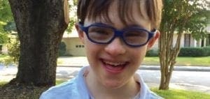 A young boy with Down syndrome and blue glasses smiles at the camera outdoors.