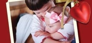 A young child holds a baby, with a red heart ornament in the corner.