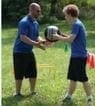 Two people in blue shirts and black shorts pass a medicine ball outdoors on a grassy field.