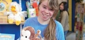 Young woman in a blue t-shirt holds a stuffed monkey in front of shelves of books and stuffed animals.