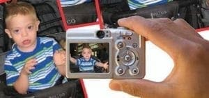 A digital camera held by a person shows a young boy in a striped shirt on its screen, with another image of the boy visible to the left.