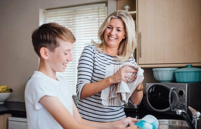A woman smiles as she dries a dish while a boy washes dishes at a sink.