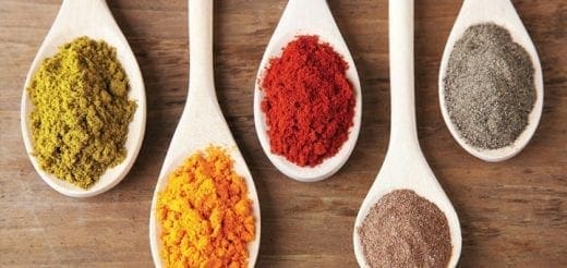 Five wooden spoons filled with colorful spices on a wooden surface.