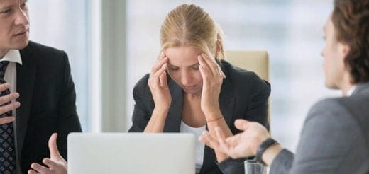 A woman in a suit rubs her temples while two men talk in a meeting.